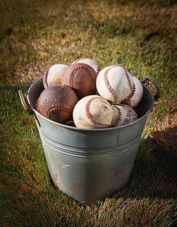 Assorted baseballs sit in a weathered bucket, surrounded by lush green grass, evoking summer play.の素材