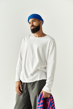 A handsome man in a white shirt and blue beanie stands confidently, holding a striped item.