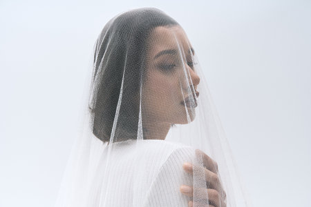 A radiant young bride stands delicately, draped in a fine veil, exuding grace and beauty.