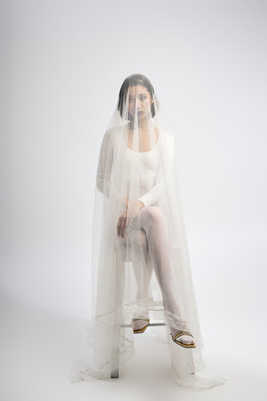Beautiful young bride sits gracefully, draped in a gossamer veil, emanating tranquility and charm.の写真素材