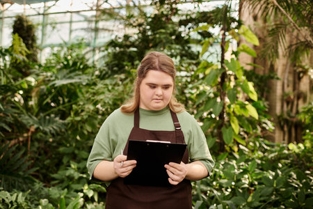 Young woman with Down syndrome shows confidence while working in a lively greenhouse.のeditorial素材