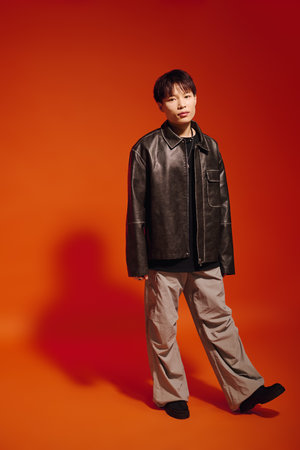 A stylish young Asian man poses confidently in casual wear against a vibrant orange backdrop.