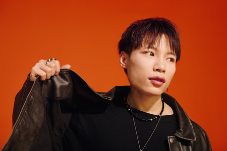 A stylish young Asian man in a leather jacket poses confidently against an orange background.のeditorial素材