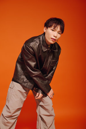 A stylish young man in a leather jacket and casual trousers stands before a bold orange backdrop.