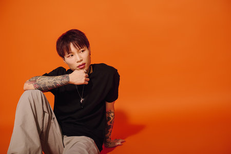 Young man with tattoos and a stylish outfit sits thoughtfully against a bold orange wall.のeditorial素材