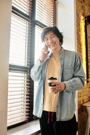 A young individual smiles while talking on the phone, holding a coffee cup in a bright office.のeditorial素材
