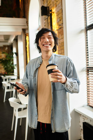 Young man smiles broadly while holding a coffee cup and phone in a lively office environment.のeditorial素材
