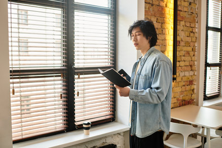 A man contemplates his thoughts while browsing a notebook by large office windows.のeditorial素材