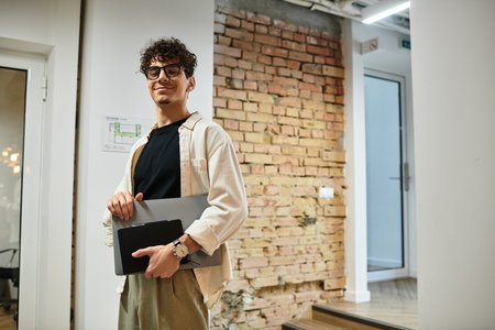 A man with curly hair holds a laptop and notebook while smiling in a stylish workspace.のeditorial素材