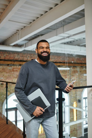 Smiling individual with a laptop and phone enjoying a moment in a contemporary workspace.のeditorial素材