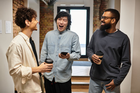 Three professionals engage in lighthearted discussion in a stylish office hallway with coffee.のeditorial素材