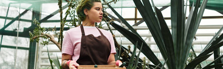 A young woman holds a wooden box in a lively greenhouse, tending to her plants with care.のeditorial素材