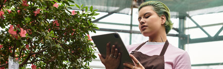 In a lush greenhouse, a young woman examines plant info on a tablet among the flowers.のeditorial素材