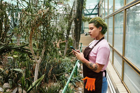 A young woman in a greenhouse checks her phone while surrounded by lush greenery and plants.のeditorial素材