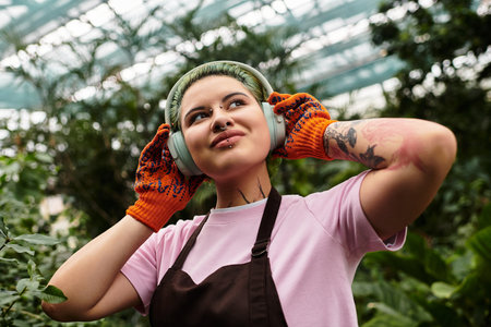 Woman with colorful hair and gloves listens to music while tending to plants in a greenhouse.のeditorial素材