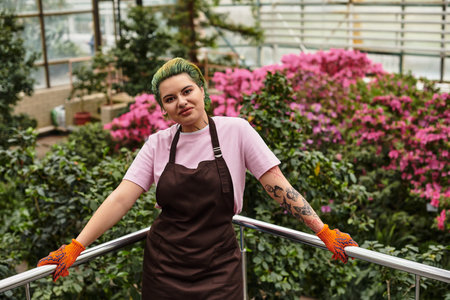 Young woman stands confidently among lush greenery and colorful flowers in a greenhouse.のeditorial素材
