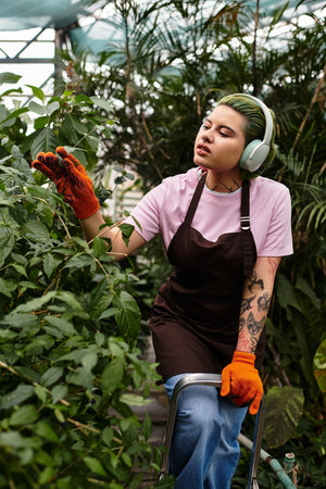 Amid lush greenery, a young woman cares for plants in a colorful greenhouse while enjoying music.のeditorial素材