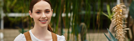Brightly lit greenhouse features a young woman smiling while surrounded by lush plants and foliage.