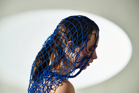 A young woman stands gracefully in a serene space, framed by vibrant blue netting.の写真素材