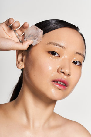 A young beautiful asian woman holds an ice cube to her glowing skin while pampering herself.の写真素材