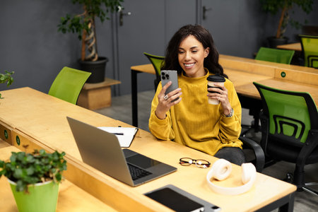 A young, elegant woman smiles at her phone while sipping coffee in a chic workspace.
