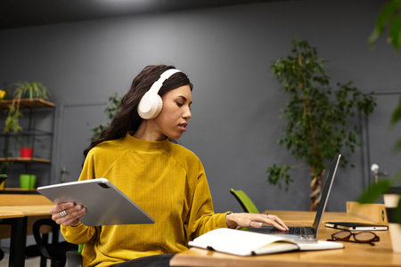 Elegant young professional focused, balancing tasks with a tablet and laptop in a chic office.