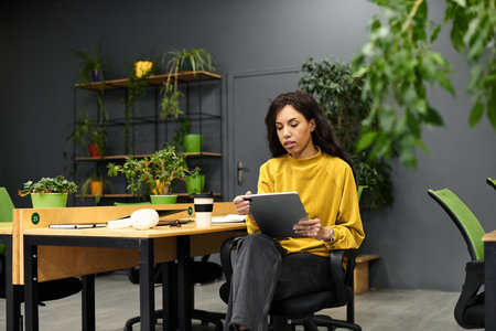 A young woman in a yellow sweater works diligently on her tablet in a plant filled office.