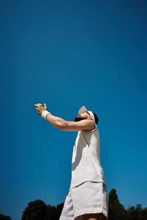 A skilled player in white sportswear prepares to serve a tennis ball under a clear blue sky.の写真素材