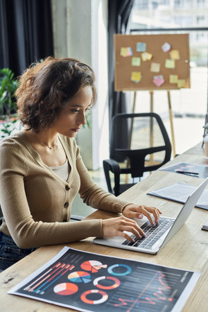 Focused individual types on a laptop, examining charts in a bright workspace.