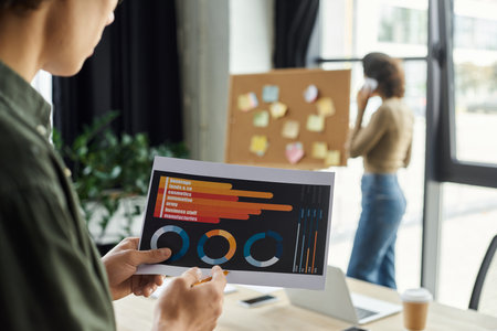 A person analyzes data charts while a colleague discusses ideas in a bright office setting.の写真素材