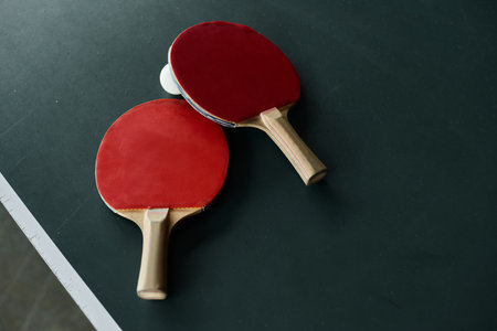 Two ping pong paddles rest beside a ball on a green table in a lively office environment.の写真素材