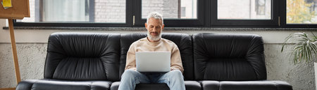 Man sits comfortably on a leather couch, intently working on a laptop in a bright office space.の写真素材
