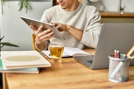 Stylish young man browses on a tablet in a cozy home office, with tea and a notebook nearby.