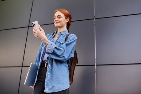 A beautiful woman with red hair stands confidently, checking her phone against a sleek backdrop.