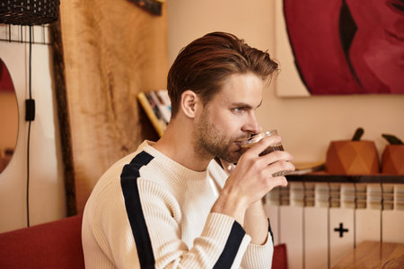 A young man with stylish hair relaxes at an inviting cafe, sipping a warm drink thoughtfully.