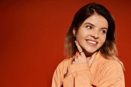 A pretty young woman showcases her stylish outfit while smiling joyfully on an orange background.
