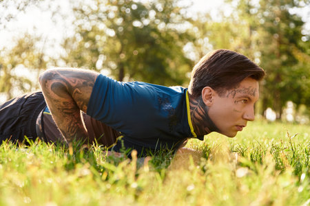 A young man with tattoos performs push ups on the grass in a sunlit park.