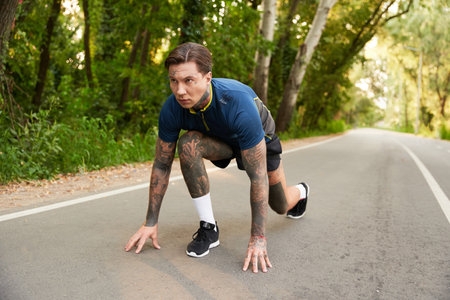 A young man with tattoos kneels, ready to start his jog on a serene road surrounded by greenery.
