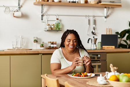 Young African American woman savors a colorful salad while seated at her stylish kitchen table.の写真素材