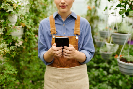 A young woman in an apron checks her phone amidst thriving plants in a greenhouse setting.の写真素材