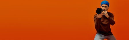 Handsome young man poses confidently with a camera against a bold orange backdrop.