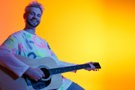 A handsome young man smiles brightly while strumming an acoustic guitar in warm hues of orange.