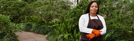 An African American woman with braids stands confidently among thriving greenery in a greenhouse,...