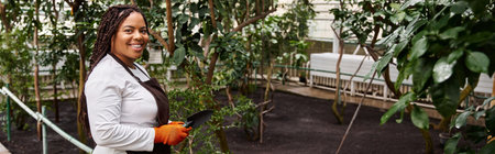 An African American woman with braids tends to plants in a lush greenhouse, cultivating her passion,...