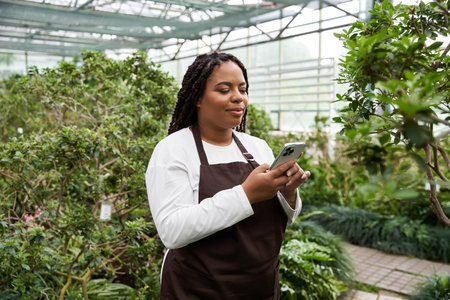 An African American woman with braids smiles while checking her phone in a lush greenhouse.