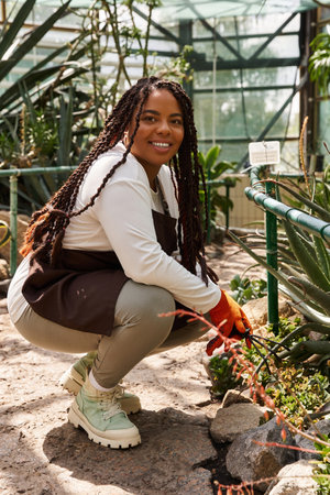 A smiling African American woman with braids tends to lush plants in a sunny greenhouse.