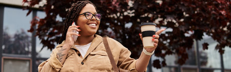 A casually dressed woman smiles, chats on her phone, and sips coffee by university buildings.のeditorial素材
