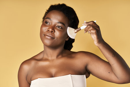Young African American woman applies makeup with a brush, showcasing her glowing skin in a studio.の写真素材