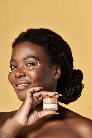 Bright-backgrounded young woman with glowing skin smiles while showing her skincare product.