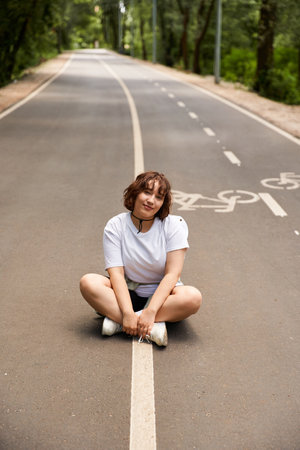 A young woman sits on a quiet road surrounded by trees, embracing the warmth of summer.の写真素材